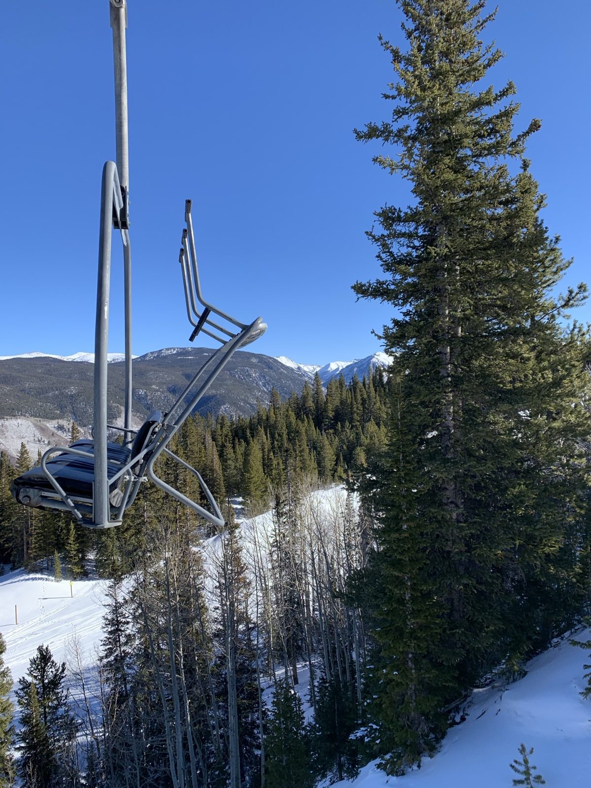 An empty chairlift chair above snowy pines on Aspen Mountain, with snow-capped peaks beyond under a clear blue sky