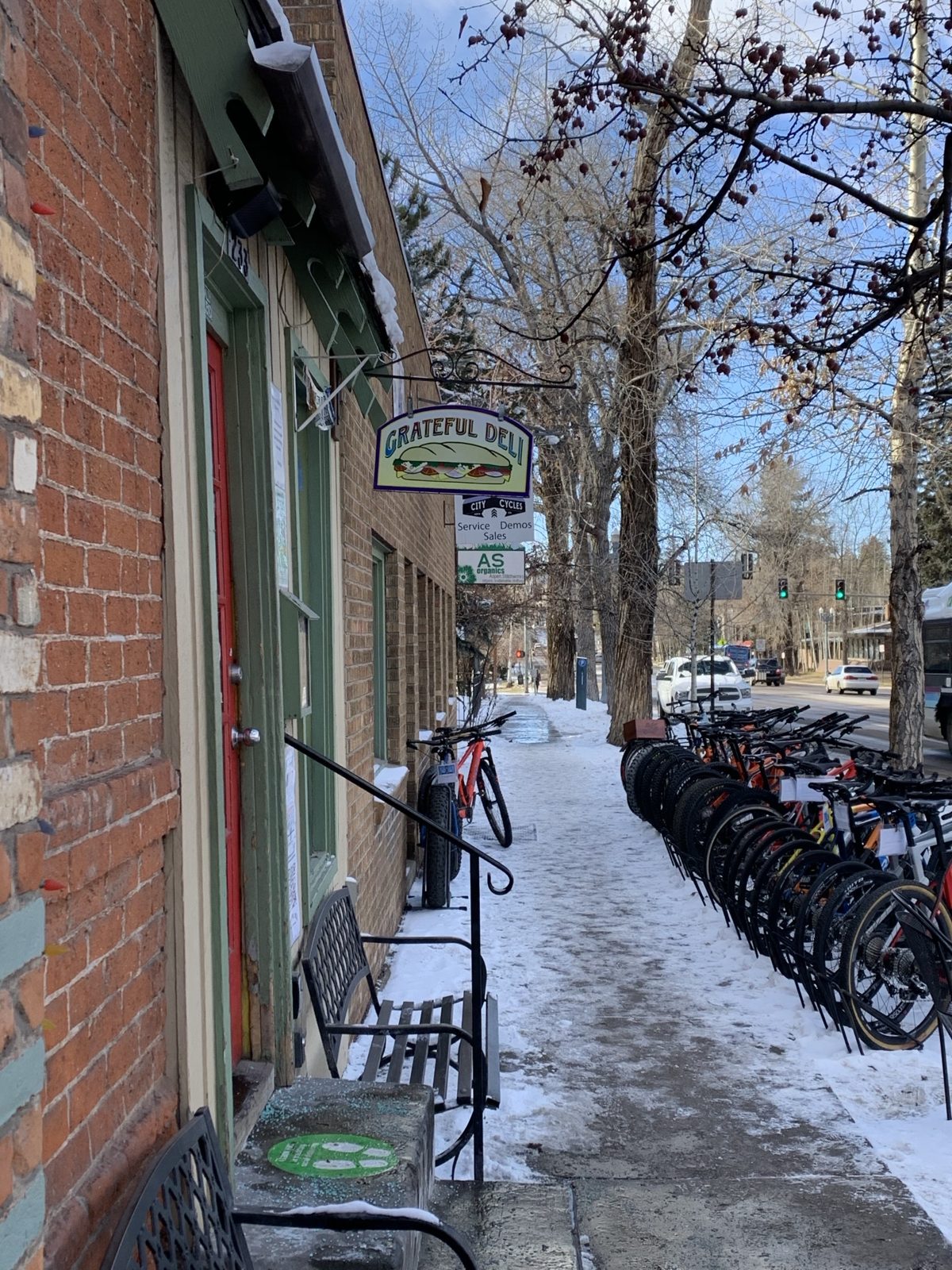 The Grateful Deli sign hanging over a snowy sidewalk on Main Street, with a rack of bikes outside