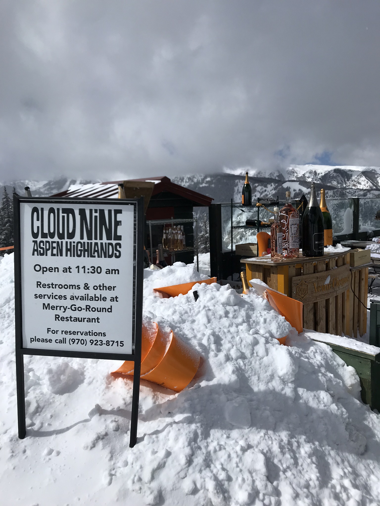 The Cloud Nine sign in deep snow at Aspen Highlands, with champagne bottles lined up on an outdoor bar