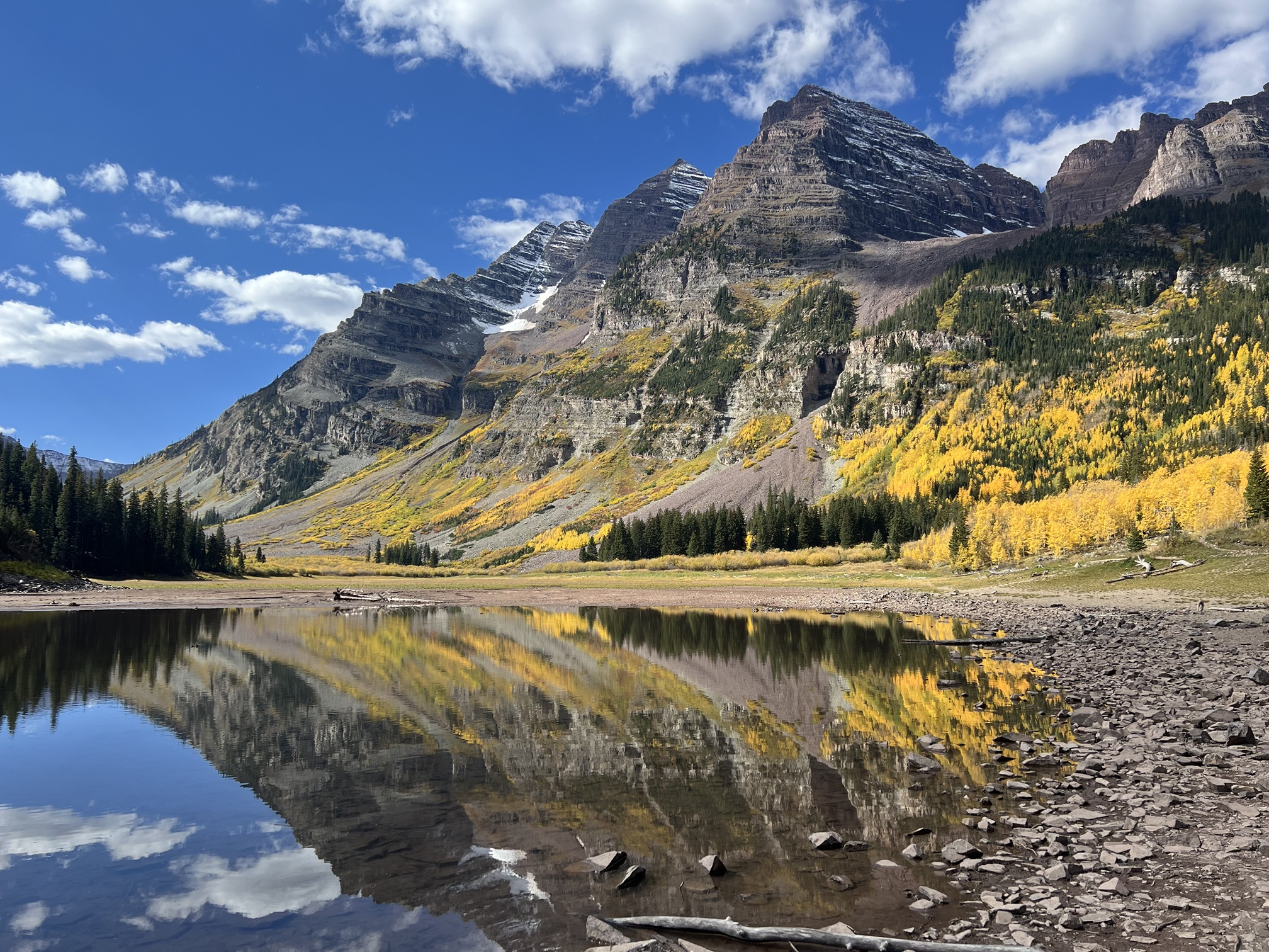 The Maroon Bells and golden aspens reflected in still water beside a rocky shoreline in fall