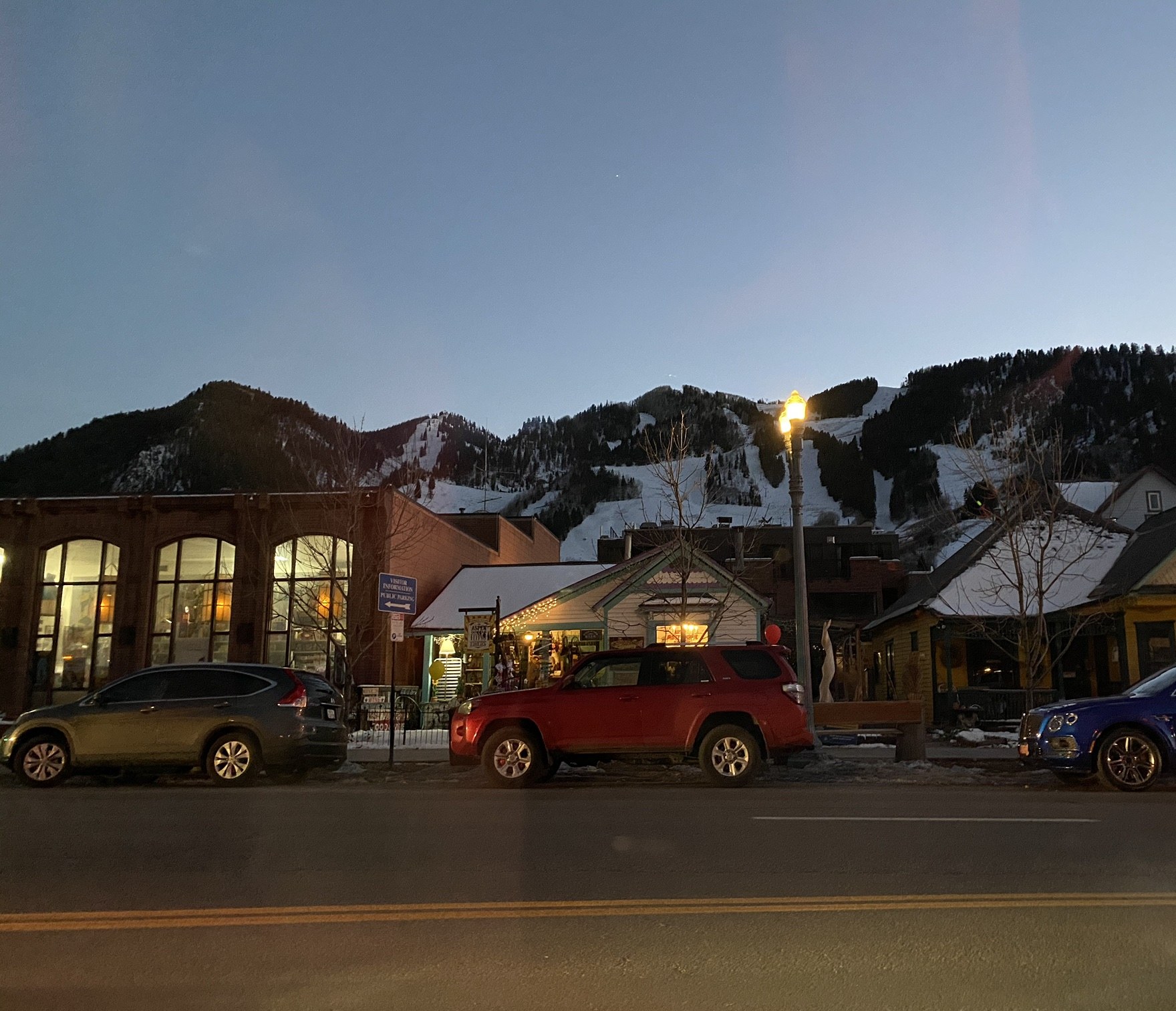 Shops and historic houses in downtown Aspen at dusk, with the snowy runs of Aspen Mountain behind
