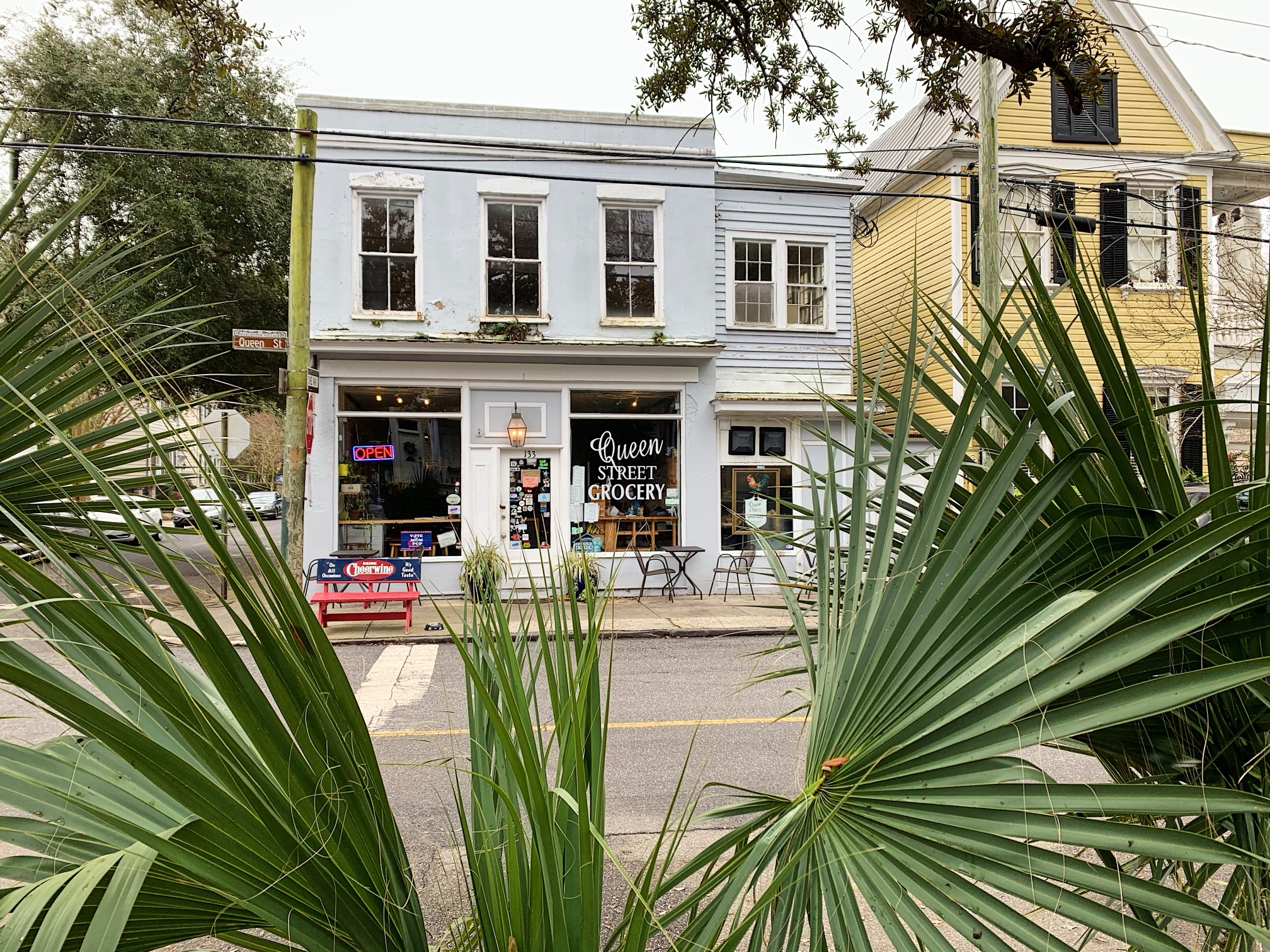 The pale blue corner building of Queen Street Grocery, framed by palmetto fronds, with tables and a red bench out front